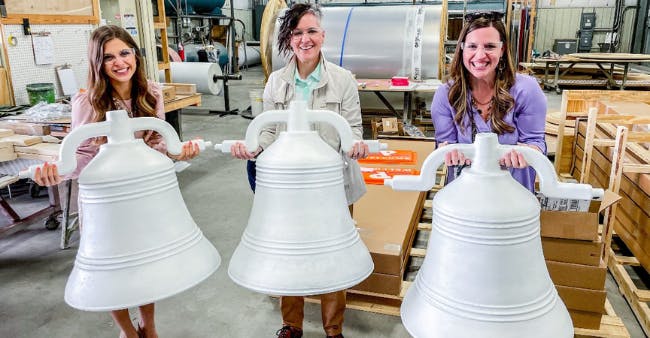 Three women smile while holding giant silver bells.